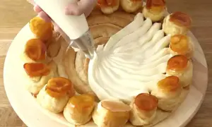 Préparation d'une recette crème chiboust Marmiton : on garnit un gâteau de crème à la poche à douille, entouré de petits choux caramélisés.
