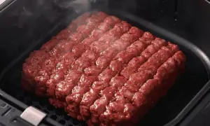 Un gros steak haché, assaisonné et prêt à cuire dans un air fryer. La vapeur s'échappe, promettant un repas savoureux.