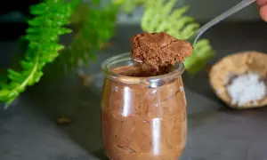 Une cuillère prélève une portion aérienne de mousse au chocolat pois chiche dans un pot en verre. Des feuilles vertes floues en arrière-plan.