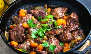 Délicieux plat de joue de porc mijotée avec des carottes et des oignons, garni de persil frais, servi dans une poêle en fonte.