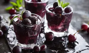 Trois verres de boisson sombre et rafraîchissante, ornés de cassis fruit et de feuilles vertes, sur un plateau noir mouillé.