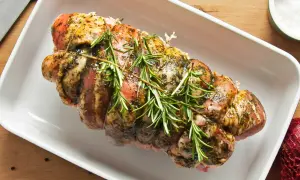 Un rôti de porc assaisonné et garni de romarin, prêt pour la cuisson au four dans un plat blanc.