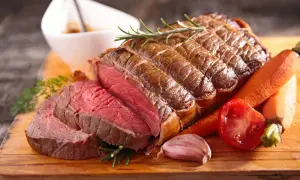 Un délicieux roti de boeuf saignant, garni de romarin, accompagné de carottes, d'ail et de tomates sur une planche en bois.