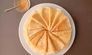 Une pile de crêpes moelleuses, pliées en éventail, sur une assiette blanche. À côté, un petit bol de sucre roux et une cuillère.