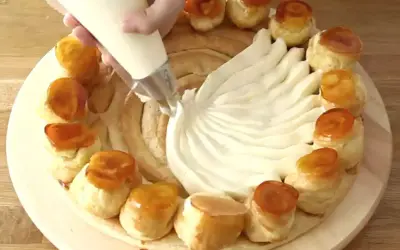 Préparation d'une recette crème chiboust Marmiton : on garnit un gâteau de crème à la poche à douille, entouré de petits choux caramélisés.