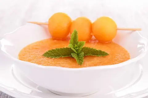 Un bol de gaspacho de melon, garni de feuilles de menthe et de billes de melon sur une brochette. Rafraîchissant et coloré.