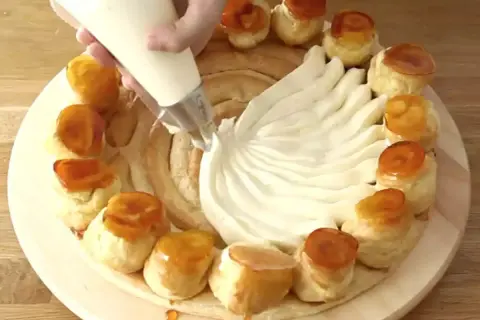 Préparation d'une recette crème chiboust Marmiton : on garnit un gâteau de crème à la poche à douille, entouré de petits choux caramélisés.
