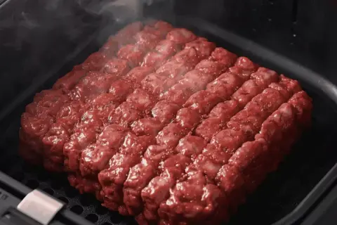 Un gros steak haché, assaisonné et prêt à cuire dans un air fryer. La vapeur s'échappe, promettant un repas savoureux.