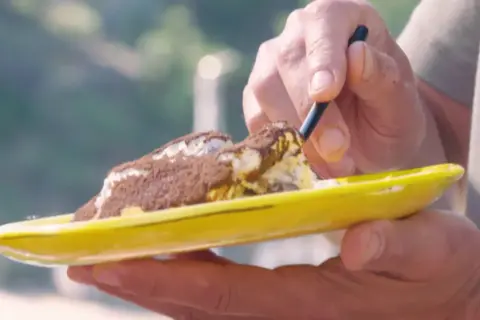 Une cuillère prélève une bouchée d'un tiramisu Laurent Mariotte, saupoudré de cacao, sur une assiette jaune vif.