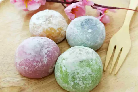 Quatre mochis colorés (rose, vert, bleu, jaune) saupoudrés de sucre glace, sur une assiette en bois avec une fleur rose. Une recette facile à réaliser !