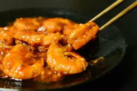 Gambas plancha enrobées d'une sauce orangée, une crevette est saisie par des baguettes.