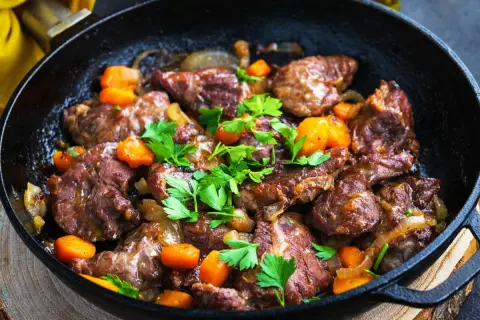 Délicieux plat de joue de porc mijotée avec des carottes et des oignons, garni de persil frais, servi dans une poêle en fonte.