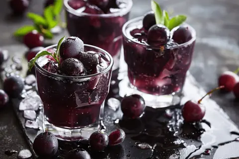 Trois verres de boisson sombre et rafraîchissante, ornés de cassis fruit et de feuilles vertes, sur un plateau noir mouillé.