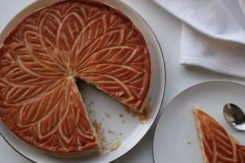 Une galette aux amandes dorée, décorée de motifs feuillus, est servie sur une assiette. Une part est déjà coupée.