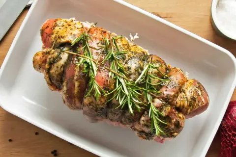 Un rôti de porc assaisonné et garni de romarin, prêt pour la cuisson au four dans un plat blanc.