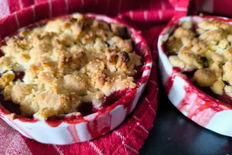 Deux petits ramequins de crumble aux quetsches, aux bords rouges et blancs, sur un torchon à rayures rouges.