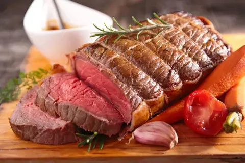 Un délicieux roti de boeuf saignant, garni de romarin, accompagné de carottes, d'ail et de tomates sur une planche en bois.