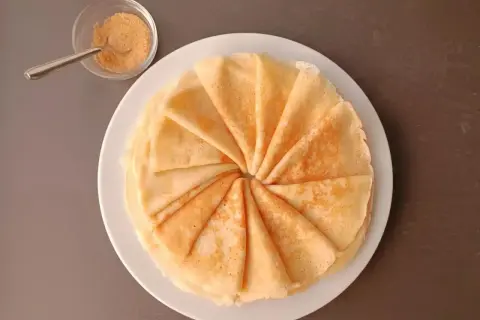 Une pile de crêpes moelleuses, pliées en éventail, sur une assiette blanche. À côté, un petit bol de sucre roux et une cuillère.