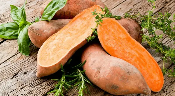 Patates douces coupées, prêtes pour la cuisson à l'eau bouillante, garnies de basilic et de thym frais sur fond de bois rustique.