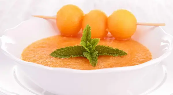 Un bol de gaspacho de melon, garni de feuilles de menthe et de billes de melon sur une brochette. Rafraîchissant et coloré.