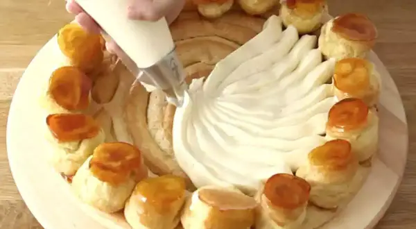 Préparation d'une recette crème chiboust Marmiton : on garnit un gâteau de crème à la poche à douille, entouré de petits choux caramélisés.