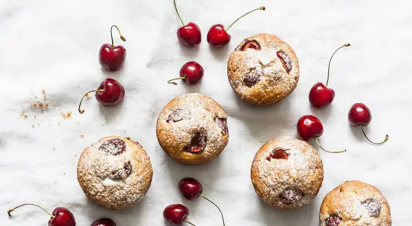 Financiers aux cerises saupoudrés de sucre glace, disposés avec des cerises fraîches sur un tissu blanc.