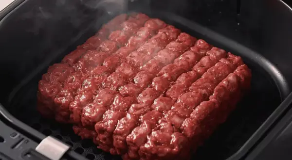 Un gros steak haché, assaisonné et prêt à cuire dans un air fryer. La vapeur s'échappe, promettant un repas savoureux.