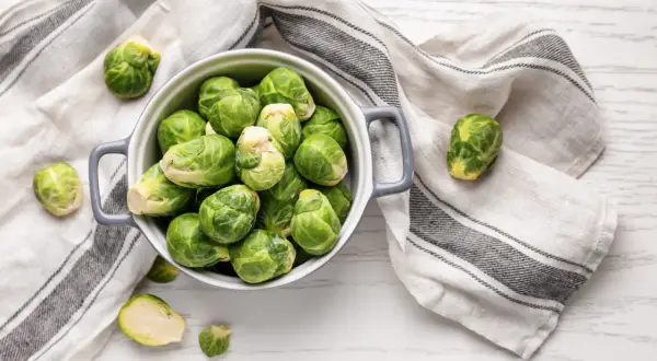Un bol gris rempli de choux de Bruxelles frais, prêts pour la cuisson. Quelques choux sont éparpillés autour, sur une serviette rayée.