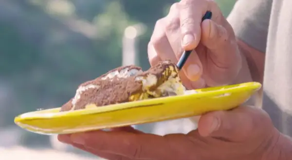 Une cuillère prélève une bouchée d'un tiramisu Laurent Mariotte, saupoudré de cacao, sur une assiette jaune vif.