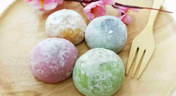 Quatre mochis colorés (rose, vert, bleu, jaune) saupoudrés de sucre glace, sur une assiette en bois avec une fleur rose. Une recette facile à réaliser !