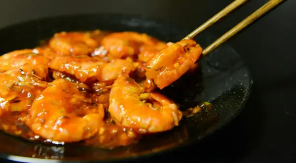 Gambas plancha enrobées d'une sauce orangée, une crevette est saisie par des baguettes.