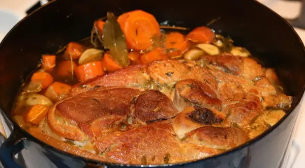 Rouelle de porc mijotant dans une cocotte avec des carottes, de l'ail et des herbes.