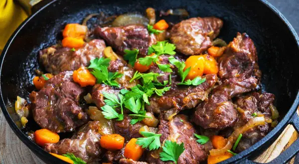 Délicieux plat de joue de porc mijotée avec des carottes et des oignons, garni de persil frais, servi dans une poêle en fonte.