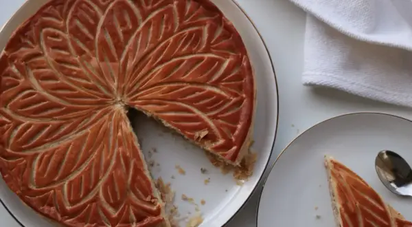 Une galette aux amandes dorée, décorée de motifs feuillus, est servie sur une assiette. Une part est déjà coupée.