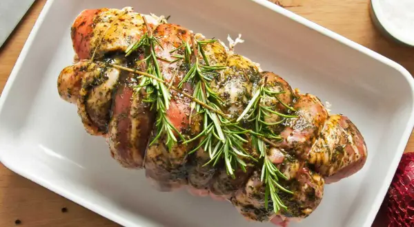 Un rôti de porc assaisonné et garni de romarin, prêt pour la cuisson au four dans un plat blanc.