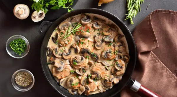 Une poêle remplie d'escalope à la crème et de champignons, garnie de persil frais et d'une branche de romarin.