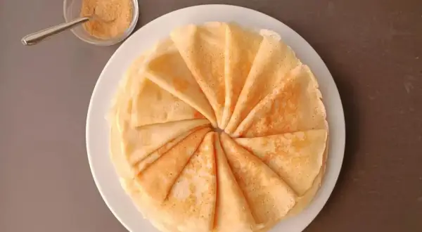 Une pile de crêpes moelleuses, pliées en éventail, sur une assiette blanche. À côté, un petit bol de sucre roux et une cuillère.
