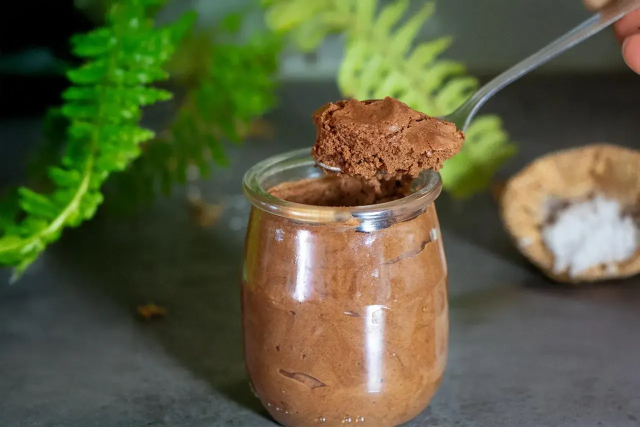 Une cuillère prélève une portion aérienne de mousse au chocolat pois chiche dans un pot en verre. Des feuilles vertes floues en arrière-plan.