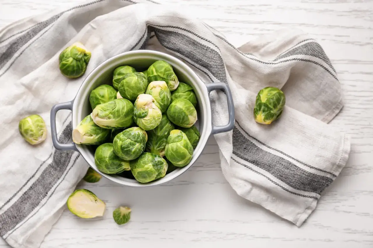 Un bol gris rempli de choux de Bruxelles frais, prêts pour la cuisson. Quelques choux sont éparpillés autour, sur une serviette rayée.