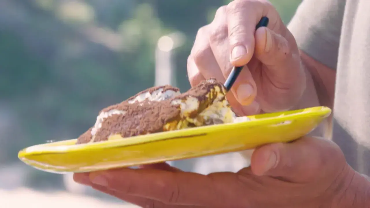 Une cuillère prélève une bouchée d'un tiramisu Laurent Mariotte, saupoudré de cacao, sur une assiette jaune vif.
