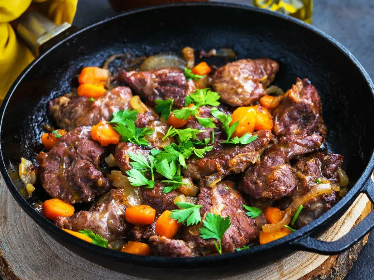 Délicieux plat de joue de porc mijotée avec des carottes et des oignons, garni de persil frais, servi dans une poêle en fonte.