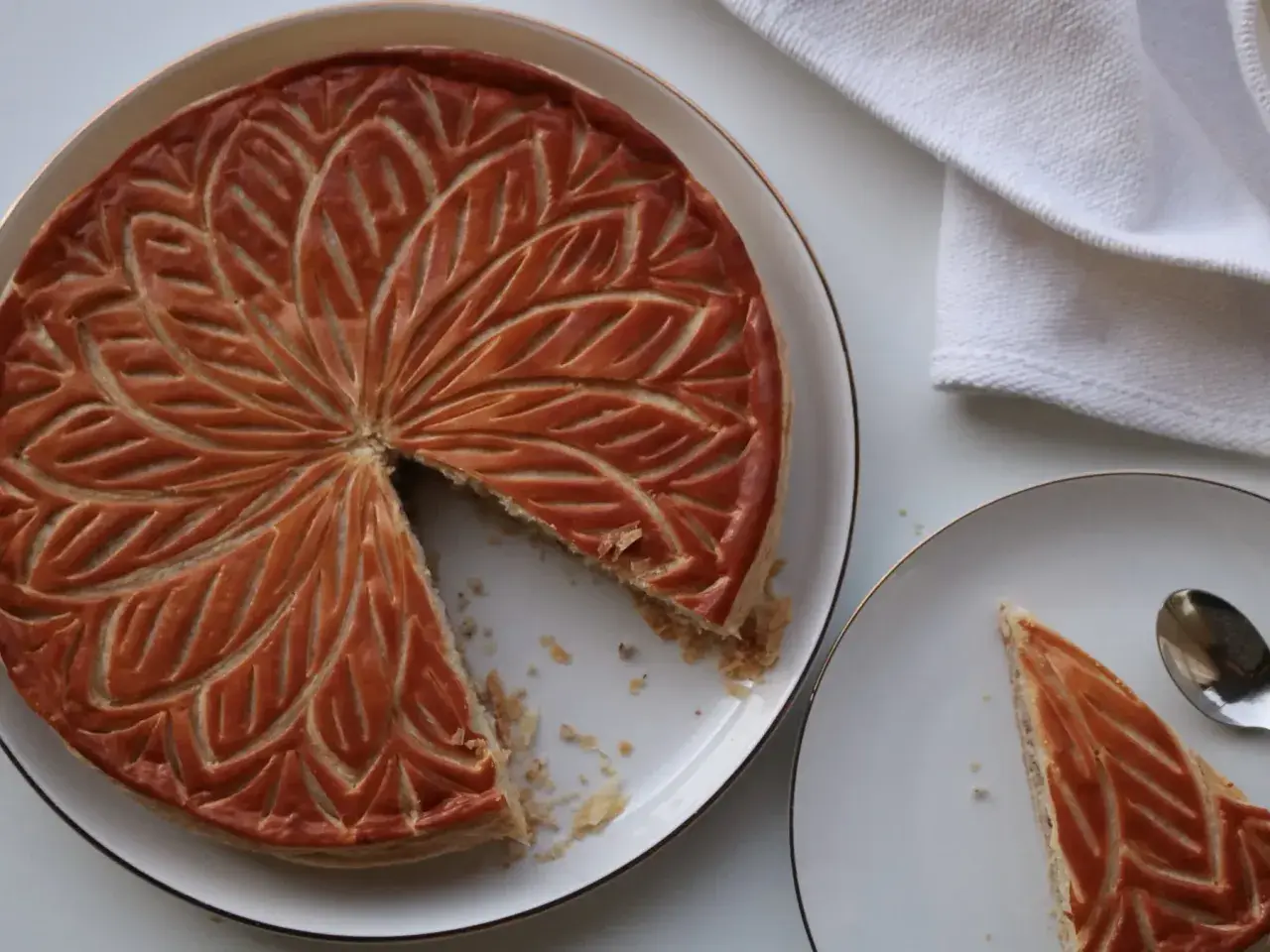 Une galette aux amandes dorée, décorée de motifs feuillus, est servie sur une assiette. Une part est déjà coupée.
