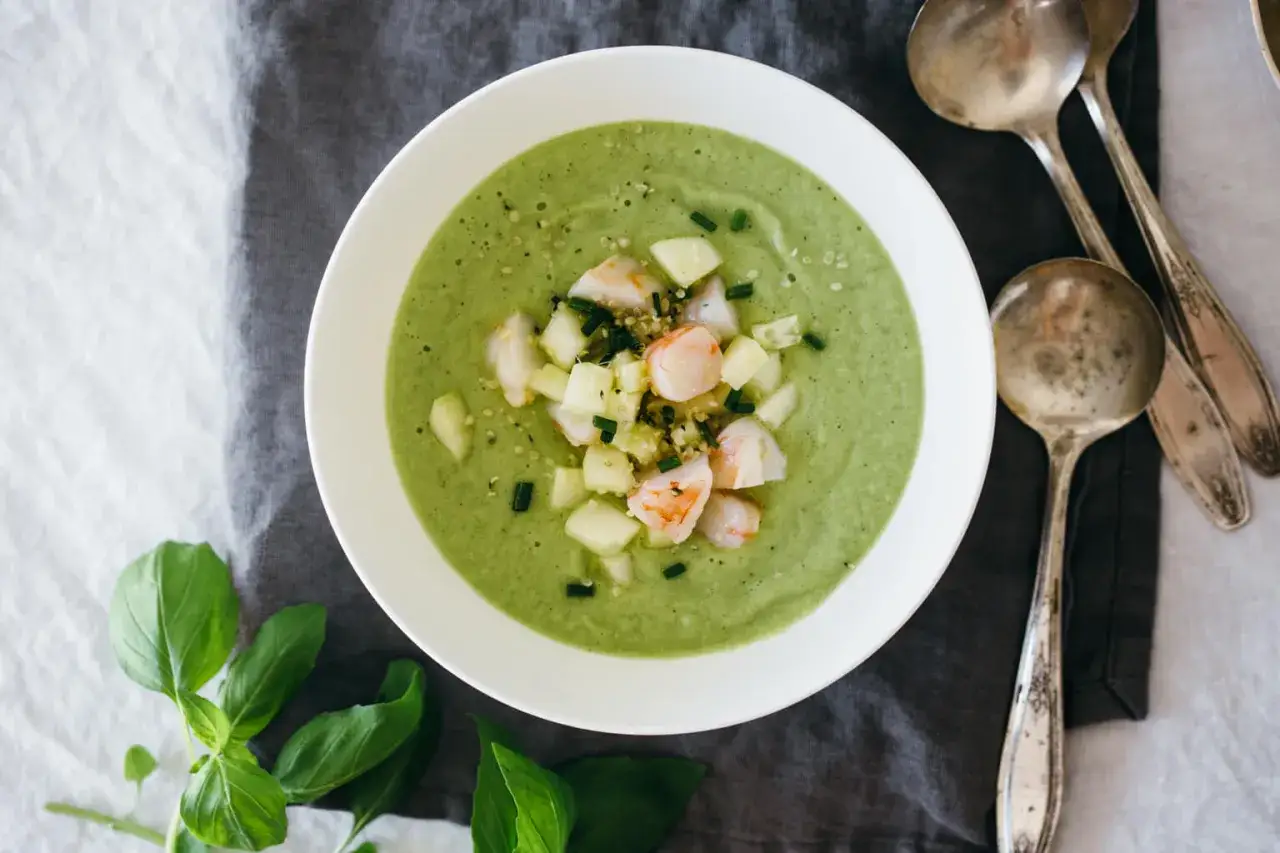 Un bol de gaspacho de melon vert, garni de crevettes et de dés de concombre, avec des cuillères anciennes à côté.