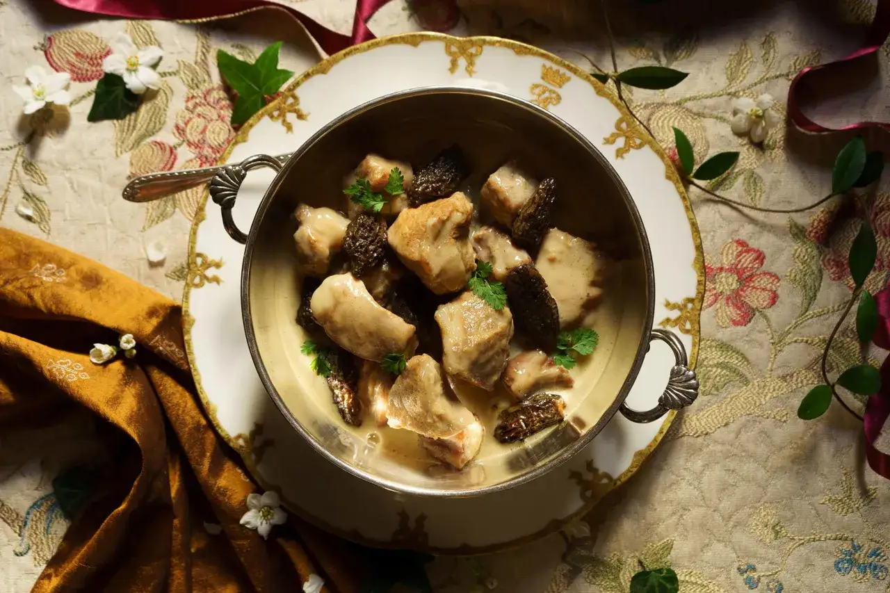 Un plat de veau aux morilles, servi dans un bol argent&eacute; sur une assiette orn&eacute;e, entour&eacute; de fleurs et de tissus luxueux.