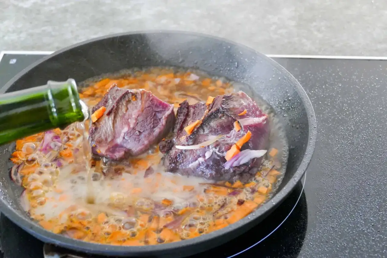 Du jus de boeuf mijote dans une poêle avec des carottes et des oignons. Une bouteille verte verse du liquide.