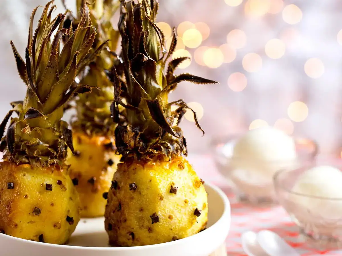 Trois petits ananas r&ocirc;tis piquet&eacute;s de clous de girofle, servis avec des boules de glace dans des coupelles, sur un fond de lumi&egrave;res douces.