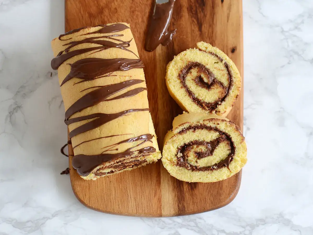 Un roulé au Nutella, entier et tranché, décoré de chocolat fondu sur une planche en bois.