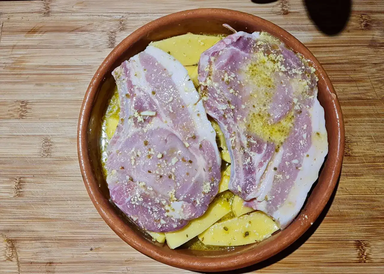 Deux c&ocirc;tes de porc crues, assaisonn&eacute;es, reposent sur des pommes de terre coup&eacute;es en tranches dans un plat en terre cuite, pr&ecirc;tes pour une recette de c&ocirc;te de porc au four.