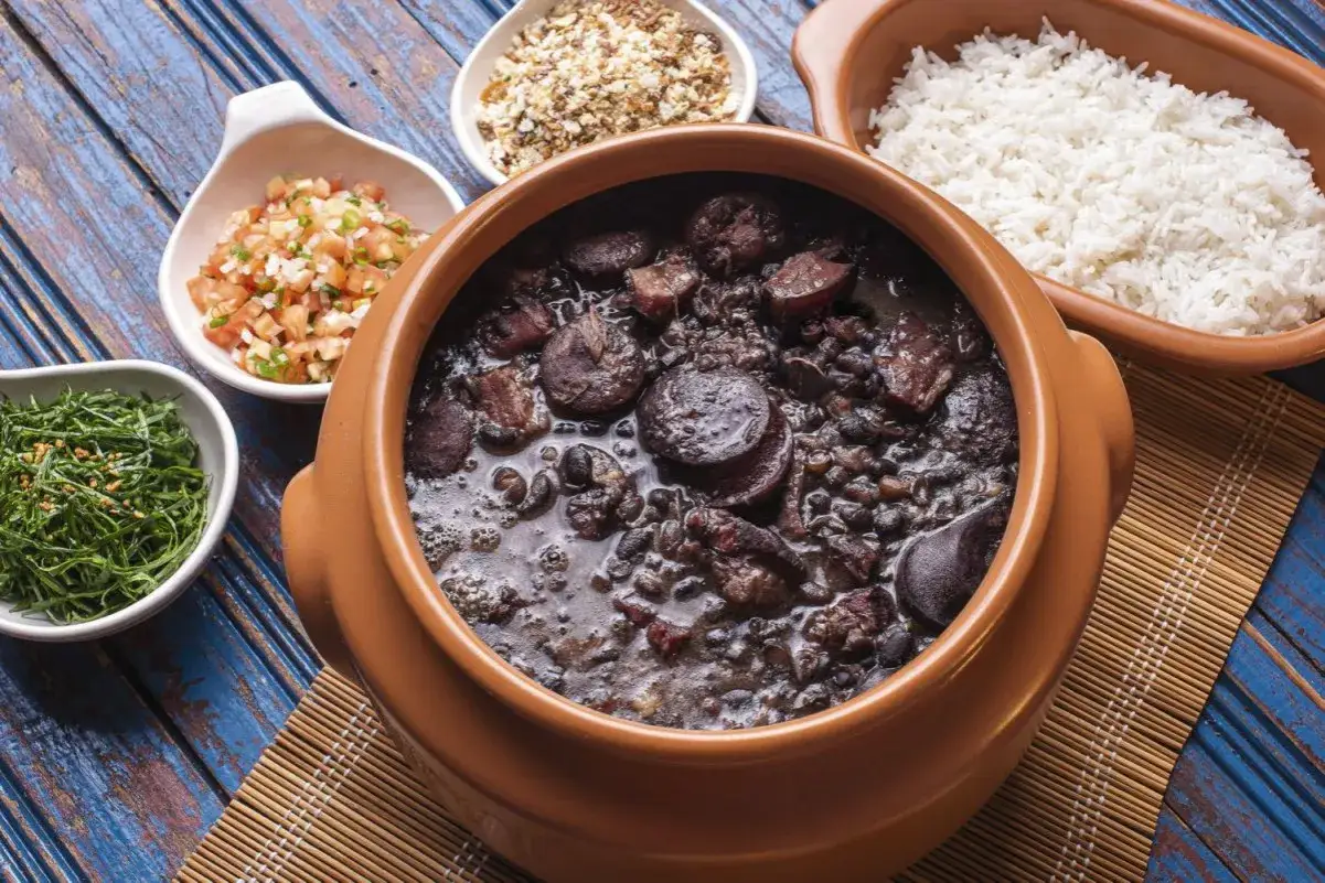 Un plat de feijoada br&eacute;silienne mijot&eacute; dans une marmite en terre cuite, accompagn&eacute; de riz blanc, de salade de tomates et de chou vert.