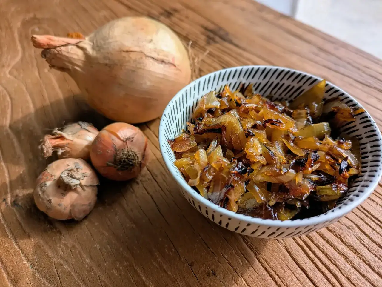 Un bol de oignons caramélisés dorés, avec des oignons crus à côté sur une planche en bois.